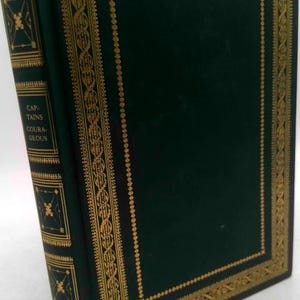 May include: A hardcover book with a dark green cover and gold decorative accents. The spine features the title "CAPTAINS COURAGEOUS" in gold lettering. The edges of the cover are also trimmed with gold.