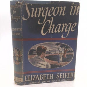 May include: A vintage hardcover book titled "Surgeon in Charge" by Elizabeth Seifert. The cover is navy blue with white lettering and a color illustration of a man and a woman near a building. The book is published by Grosset & Dunlap.