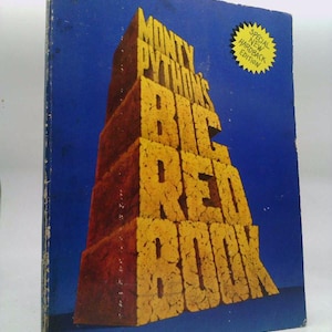May include: A vintage hardcover book titled "Monty Python's Big Red Book" with a blue cover. The title is in large, yellow, textured letters. A yellow starburst sticker reads "Special New Hardback Edition". The book is from Warner Books.