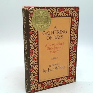May include: A hardcover book titled "A Gathering of Days" by Joan W. Blos. The cover features a tan background with the title in large letters, framed by a decorative border of leaves and red berries. A gold seal is affixed to the top.