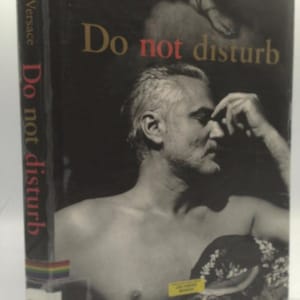 May include: A book cover featuring a black and white image of a man with the title "Do not disturb" in gold and red lettering. The author's name, Gianni Versace, is at the top. The spine displays the title in red.