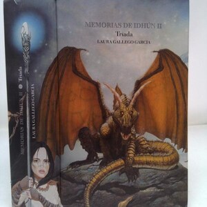 May include: A book cover featuring a detailed illustration of a dragon with orange wings and a long tail, set against a rocky landscape. The title "MEMORIAS DE IDHÚN II Triada" and author's name "LAURA GALLEGO GARCIA" are displayed.