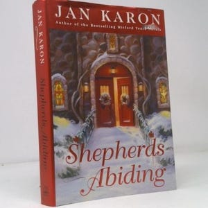 May include: A hardcover book titled "Shepherds Abiding" by Jan Karon. The cover features a detailed illustration of a church entrance with a snowy landscape. The spine is red with the author's name and title.