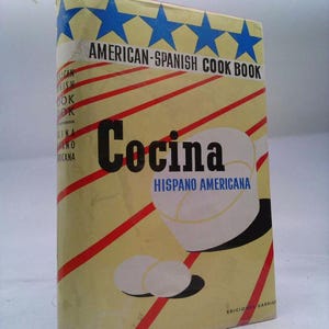 May include: A vintage cookbook titled "American-Spanish Cook Book." The cover is a light yellow with red diagonal stripes and blue stars. The word "Cocina" is prominently displayed, with the text "Hispano Americana" below it.