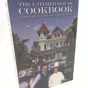 May include: The Latimer House Cookbook: From Our Kitchen to Yours, a cookbook with a photo of a couple standing in front of a large house with a blue and white exterior. The woman is wearing a white chef's hat and a white shirt. The man is wearing a blue shirt. The book is by Jean Fetzer Walters.