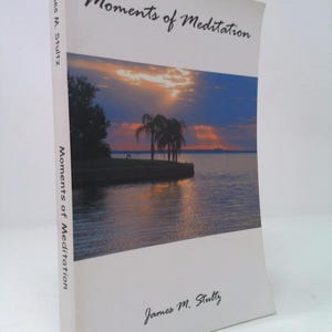 May include: A hardcover book titled "Moments of Meditation" by James M. Stultz. The cover features a scenic photograph of a sunset over water with palm trees. The book's spine also displays the title and author's name.