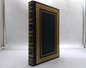 May include: A hardcover book with a black cover and gold decorative trim. The spine features gold lettering and decorative elements. The book's title is "The Odyssey".