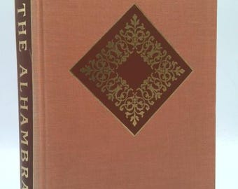 May include: A hardcover book titled "The Alhambra" with a peach-colored cover. The cover features a dark brown diamond shape with gold filigree and a smaller brown square in the center. The book's spine also has the title in gold.
