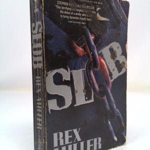 May include: A paperback book titled "Slob" by Rex Miller. The cover features a large, stylized word "Slob" in white letters against a dark background. A chain is wrapped around the letters, with a red and blue color scheme.