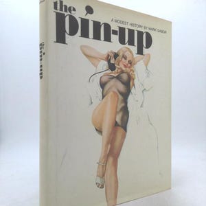 May include: A vintage book titled "The Pin-up: A Modest History by Mark Gabor." The cover features a retro illustration of a woman in lingerie holding a telephone receiver. The book has a cream-colored cover with black text.