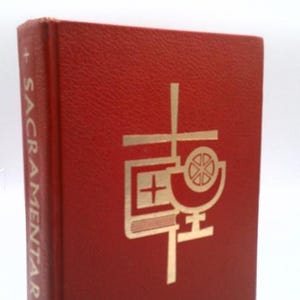 May include: A red book with gold lettering and a religious symbol on the cover. The spine reads "SACRAMENTARY" in white. The book has a textured surface and a cream-colored ribbon bookmark.