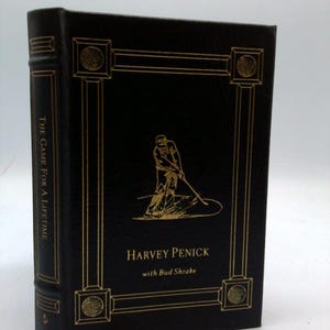 May include: A hardcover book titled "The Game For a Lifetime" by Harvey Penick with Bud Shrake. The book has a black cover with gold lettering and a gold illustration of a golfer. A gold ribbon bookmark is visible.