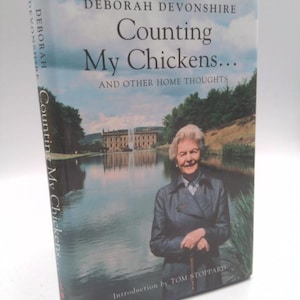May include: A book titled "Counting My Chickens..." by Deborah Devonshire. The cover features a woman standing in front of a lake with a large building in the background. The book's spine also displays the title and author.
