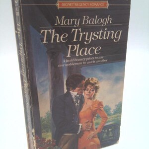 May include: A mass market paperback book titled The Trysting Place by Mary Balogh. The cover features an illustration of a man in a dark suit and a person in an orange dress standing near a stone pillar. The text reads Signet Regency Romance.