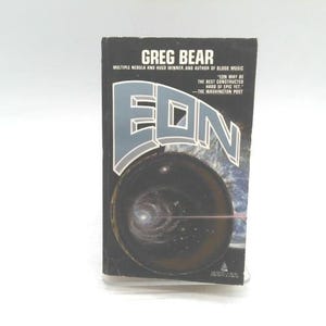 May include: A paperback book titled "Eon" by Greg Bear. The cover features a dark, circular image with a galaxy-like design, and the title is in large, stylized letters. The author's name and accolades are printed at the top.