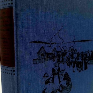 May include: A vintage book with a blue cover and a dark red spine. The cover features a black illustration of a colonial scene with buildings and figures. The spine has text and star accents.