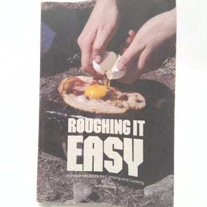 May include: A vintage cookbook titled "Roughing It Easy: A Unique Idea Book for Camping and Cooking" by Dian Thomas. The cover features a photo of a person cracking an egg over bacon cooking on a cast iron skillet.