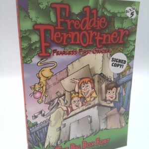 May include: A softcover book titled Freddie Fernortner Fearless First Grader Book 5 The Big Box Fort by Johnathan Rand. The cover features a cartoon illustration of three children in a cardboard box fort with a cat, and a white Signed Copy sticker.