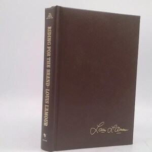 May include: A hardcover book titled "Riding for the Brand" by Louis L'Amour. The book has a dark brown cover with the title and author's name printed vertically on the spine. The author's signature is in gold on the cover.