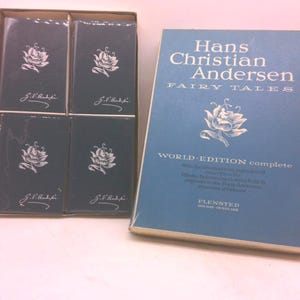 May include: A collection of Hans Christian Andersen's Fairy Tales books. The main book is blue with white text and a floral illustration. The set includes smaller, dark blue books with a floral design and the text "J.S. Bach".