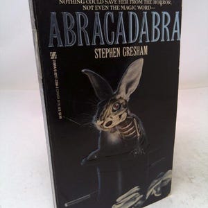 May include: A mass market paperback book titled Abracadabra by Stephen Gresham with a black cover featuring a skeletal rabbit emerging from a black top hat. The text reads: Nothing could save her from the horror. Not even the magic word—Abracadabra.