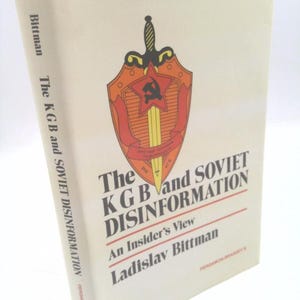 May include: A hardcover book titled "The KGB and Soviet Disinformation" by Ladislav Bittman. The cover features a shield emblem with a sword and a red star, set against a cream-colored background. The book's spine displays the author's name.