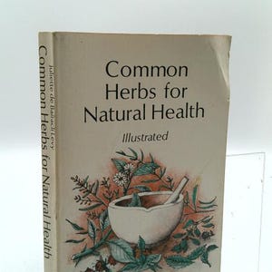 May include: A softcover book titled Common Herbs for Natural Health by Juliette de Bairacli Levy. The off-white cover features a central illustration of a mortar and pestle surrounded by various green herbs and small white flowers.