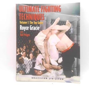 May include: A book titled Ultimate Fighting Techniques Volume 1: The Top Game by Royce Gracie with Kid Peligro. The cover features a photograph of two individuals in a Brazilian Jiu-Jitsu match inside a cage, with one person in a white gi.