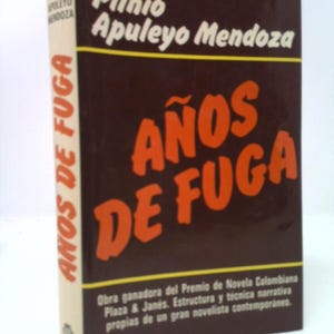 May include: A hardcover book titled "Años de Fuga" by Plinio Apuleyo Mendoza. The cover is dark brown with orange lettering. The spine is off-white with the title in vertical orange letters. The book is a winner of the Colombian Novel Prize.