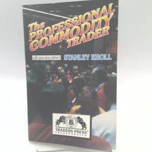 May include: A book titled The Professional Commodity Trader by Stanley Kroll. The cover features a dark, crowded trading floor scene with people in business attire. A white rectangular logo for Traders Press Incorporated is at the bottom.