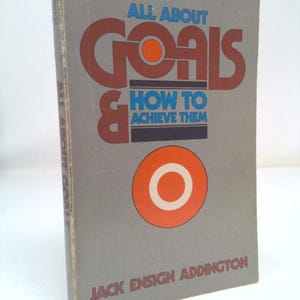 May include: A gray book with the title "All About Goals & How to Achieve Them" in colorful, bold letters. The author is Jack Ensign Addington. The cover features a large red circle with a white ring around it.