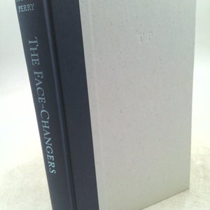 May include: A hardcover book titled "The Face-Changers" by Thomas Perry. The spine is dark blue with white text. The cover is light gray with the initials "TP" embossed on the front.