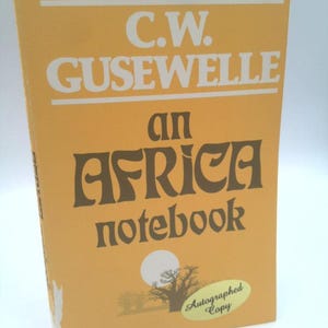 May include: A hardcover book titled "an AFRICA notebook" by C.W. GUSEWELLE. The cover is a light orange color with brown text and a graphic of a tree and sun. An oval sticker reads "Autographed Copy".