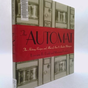May include: The cover of a red hardcover book titled "The Automat: The History, Recipes, and Allure of Horn & Hardart's Masterpiece" by Lorraine B. Diehl and Marianne Hardart. The cover features a black and white illustration of a row of vintage automats.