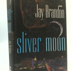 May include: A hardcover book titled "Sliver Moon" by Jay Brandon. The cover features a dark background with a crescent moon and a city skyline at the bottom. The title is in light blue, and the author's name is in white.
