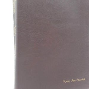 May include: A brown leather-bound book with the text "Kathy Ann Churchill" in gold lettering. The book has a textured surface and a slightly curved shape, suggesting it is well-used.