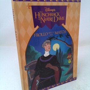 May include: A hardcover book titled "The Hunchback of Notre Dame" by Disney. The cover features an illustration of a character in a black robe with a purple collar, set against a nighttime scene. The book's spine is a light orange color with the text "Frollo Meets His Match".