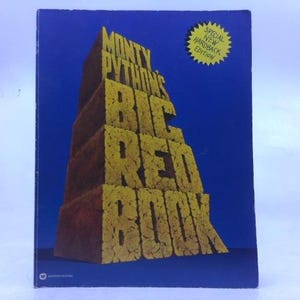 May include: A hardcover book titled "Monty Python's Big Red Book" against a blue background. The title is in yellow, block-style letters. A yellow starburst sticker reads "Special New Hardback Edition". The book has a brick-like design.