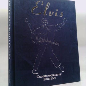 May include: A navy blue hardcover book titled "Elvis" with a silver illustration of Elvis Presley playing a guitar. The words "Commemorative Edition" are printed below the illustration. A small bookmark rests at the bottom.