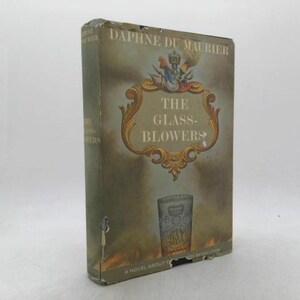 May include: A vintage hardcover book titled "The Glass-Blowers" by Daphne du Maurier. The cover features an ornate gold frame with the title, a glass illustration, and a crest. The book has a worn, aged appearance with a faded color palette.