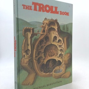 May include: A hardcover book titled "THE TROLL BOOK" by Michael Berenstain. The cover features a detailed illustration of a troll-shaped structure with internal rooms and figures, set against a green and blue backdrop.