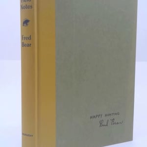 May include: A hardcover book titled "Fred Bear's Field Notes" with a bear illustration. The spine is yellow, and the cover is a muted green. The words "Happy Hunting" and a signature are on the back cover.
