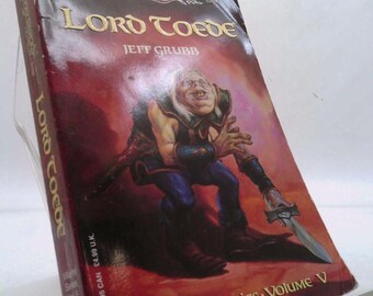 May include: A hardcover book titled "Lord Toede" by Jeff Grubb, part of the Dragonlance series. The cover features a fantasy illustration of a character holding a sword, set against a red and brown background. The book is part of the Villains Series, Volume V.