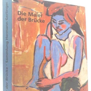 May include: A book titled "Die Maler der Brücke" with a painting on the cover. The artwork depicts a seated figure with blue skin, red accents, and a red head covering. The spine is blue with white text.