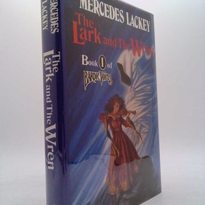 May include: A blue hardcover book with the title "The Lark and The Wren" by Mercedes Lackey. The book is part of the "Bardic Voices" series and is book 1 of the series. The cover features a woman playing a violin in front of a white bird with a blue background.