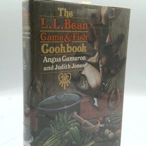 May include: The L.L. Bean Game & Fish Cookbook by Angus Cameron and Judith Jones. The hardcover book features a wood-grain background with illustrations of hanging game birds, sausages, a black pot, fresh vegetables, and a fish on the cover.