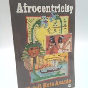 May include: A softcover book titled Afrocentricity by Molefi Kete Asante. The dark brown cover features a colorful illustration of an Egyptian pharaoh, a sun, hieroglyphics, two figures playing instruments, and a boat on the water.