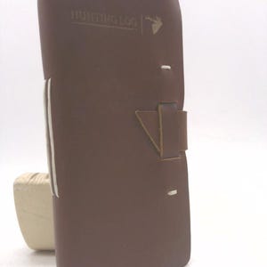 May include: A brown leather Hunting Log journal with a buck silhouette. The journal has a leather strap closure and white stitching along the spine. The journal is closed and upright.