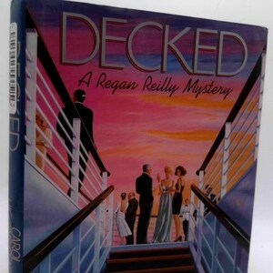 May include: A hardcover book titled "Decked: A Regan Reilly Mystery" with a cover illustration of people on a cruise ship deck at sunset. The title is in large, silver letters.