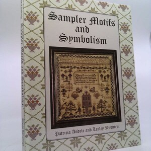 May include: A book cover with a beige background and a floral pattern. The title is "Sampler Motifs and Symbolism" with a black and white sampler image in the center. The authors are Patricia Andrle and Lesley Rudnicki.